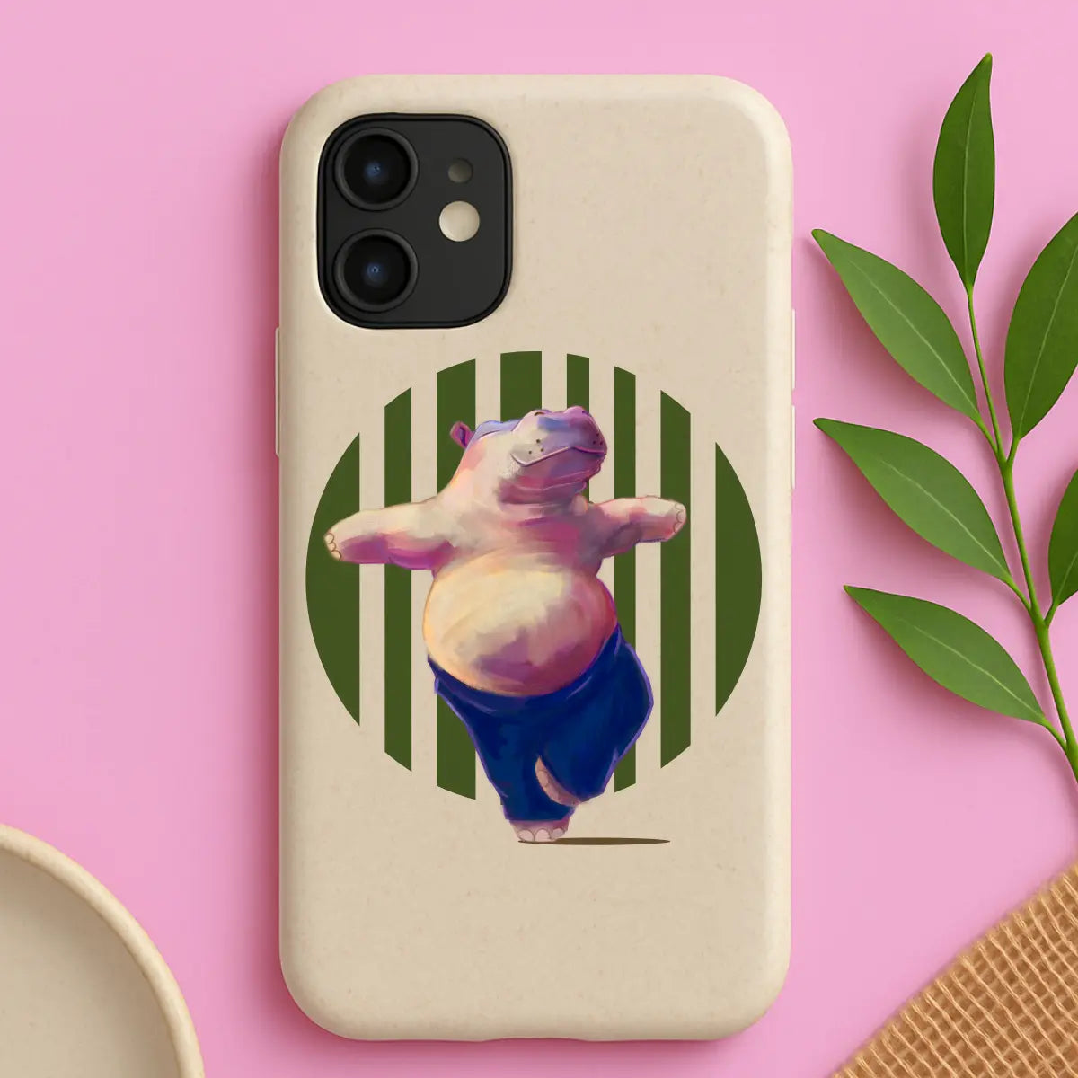 Eco-friendly illustrated yoga hippo phone case on soft pink background, styled with plant leaves — sustainable phone accessory in a lifestyle setting.

